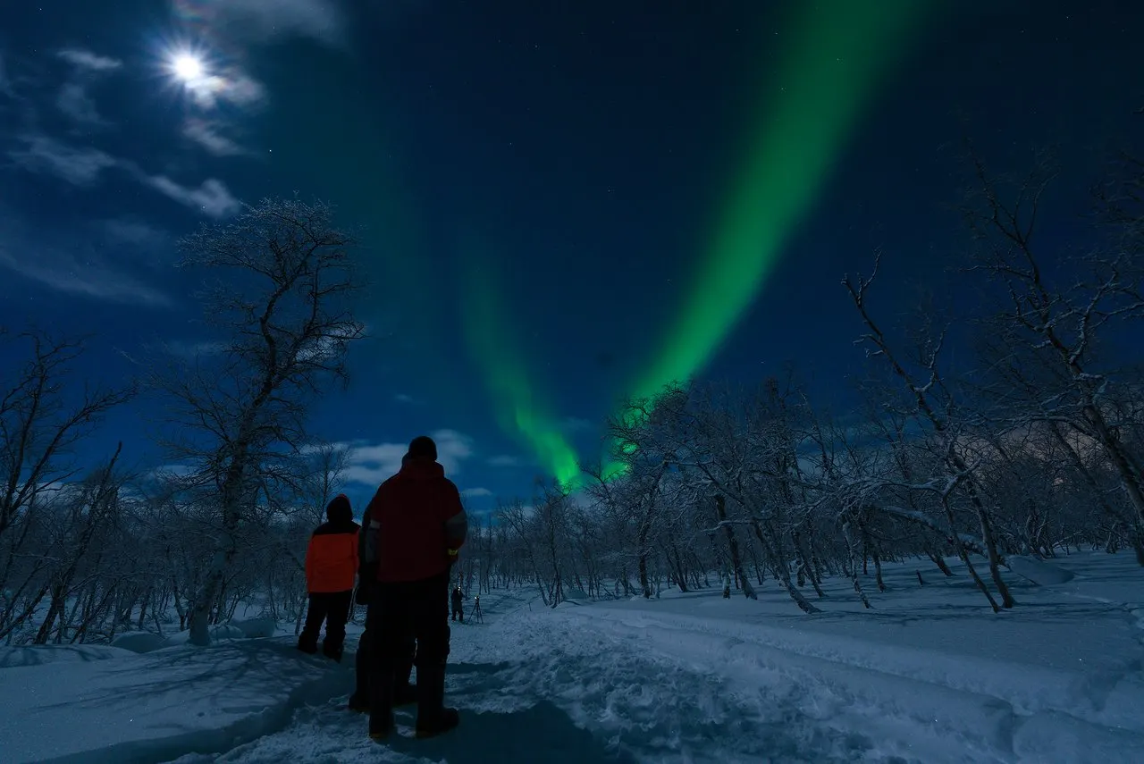 Two people in winter clothes watching a green aurora from a snowy path