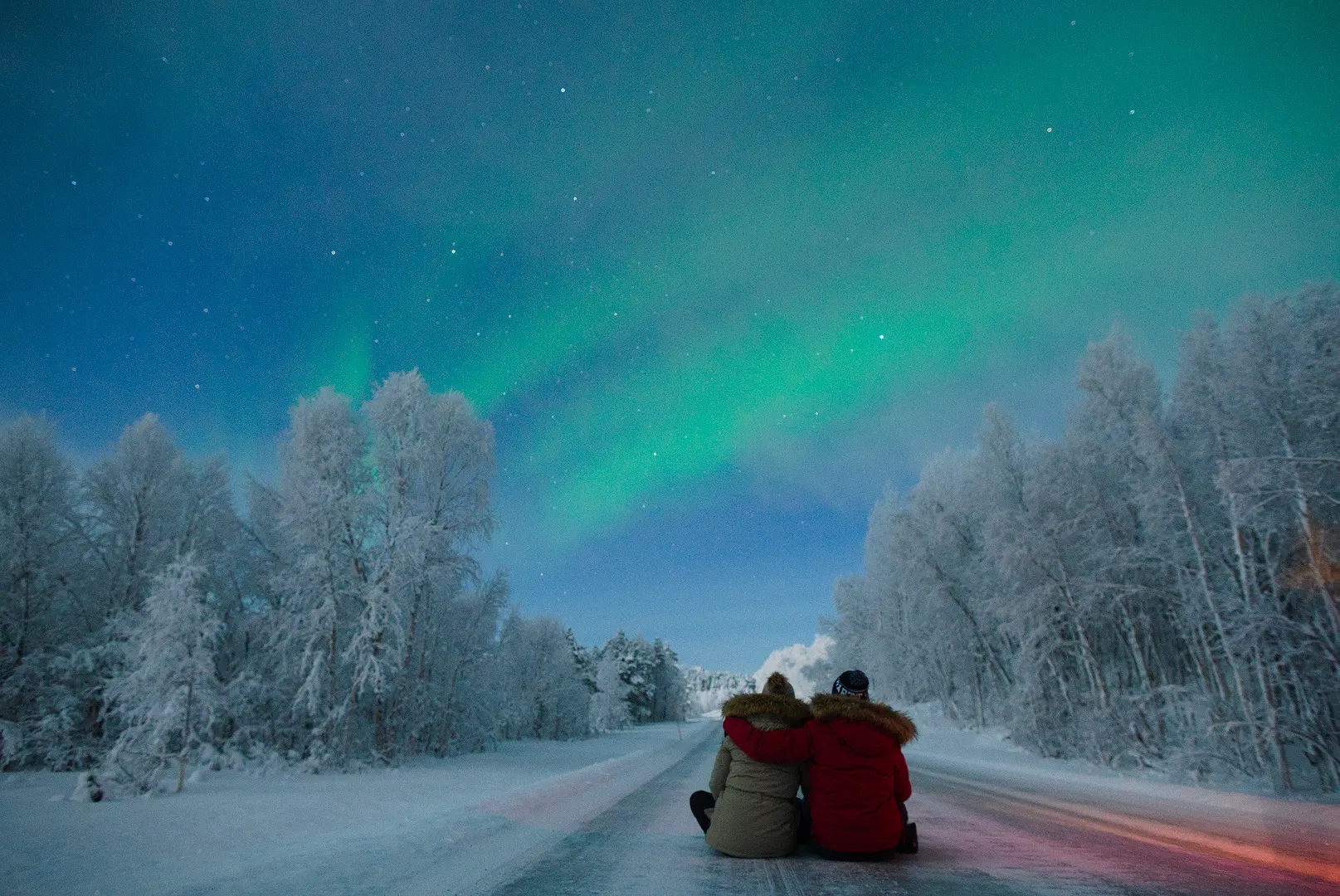Two guests in thermal suits watching the northern lights over a frosted forest road